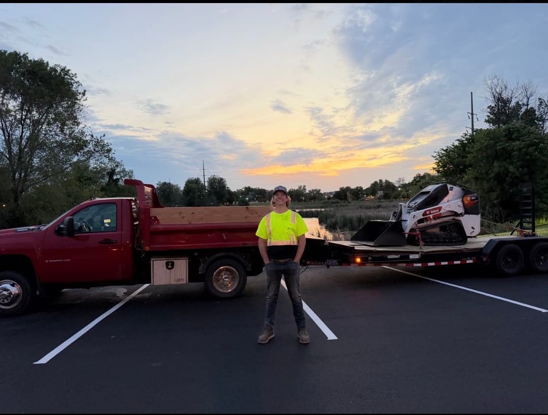 Construction worker in neon vest stands by flatbed truck at sunset with snowmobile trailer.