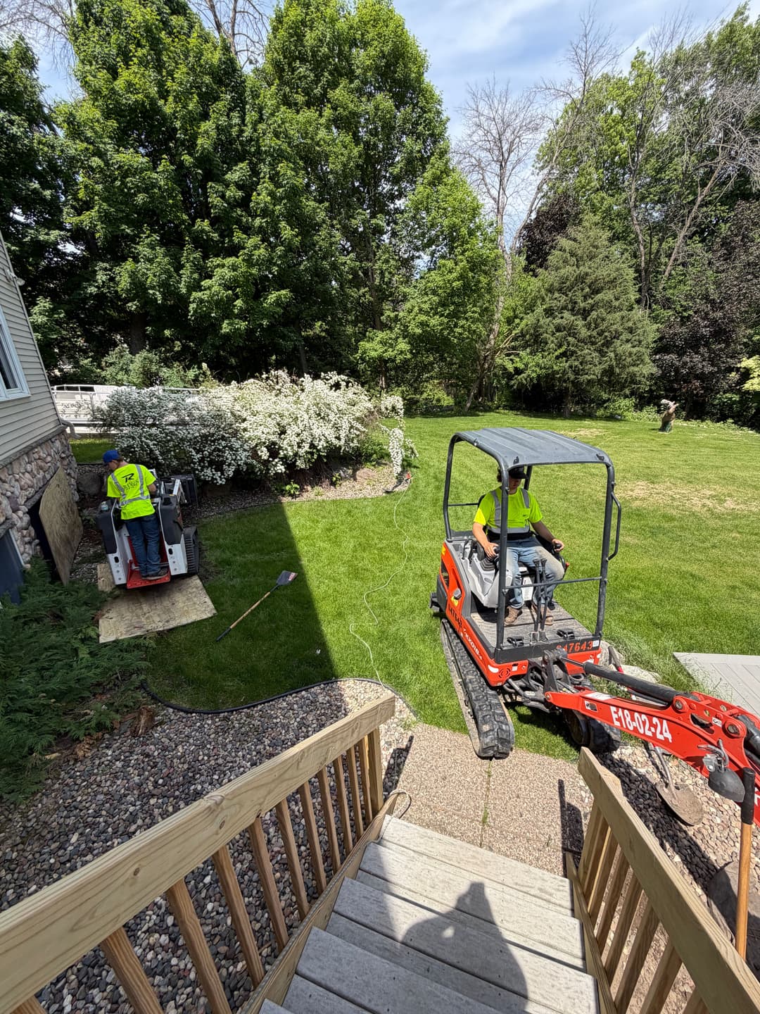 Excavator and worker on residential landscaping project in backyard with greenery.