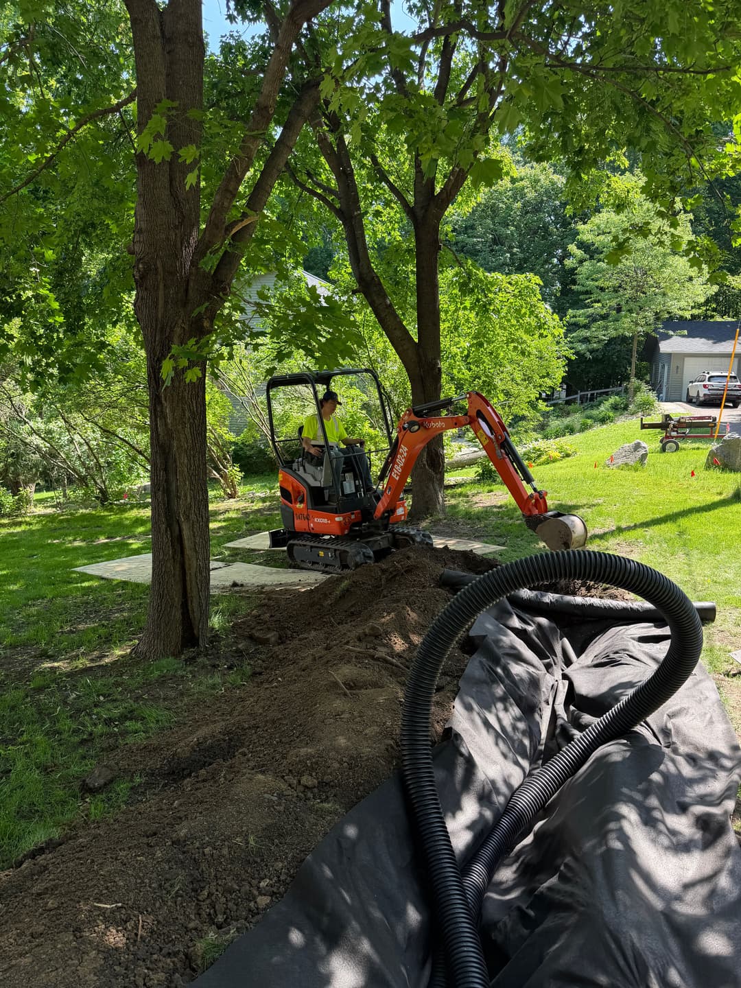Excavator digging a trench in a green yard surrounded by trees and landscaping equipment.