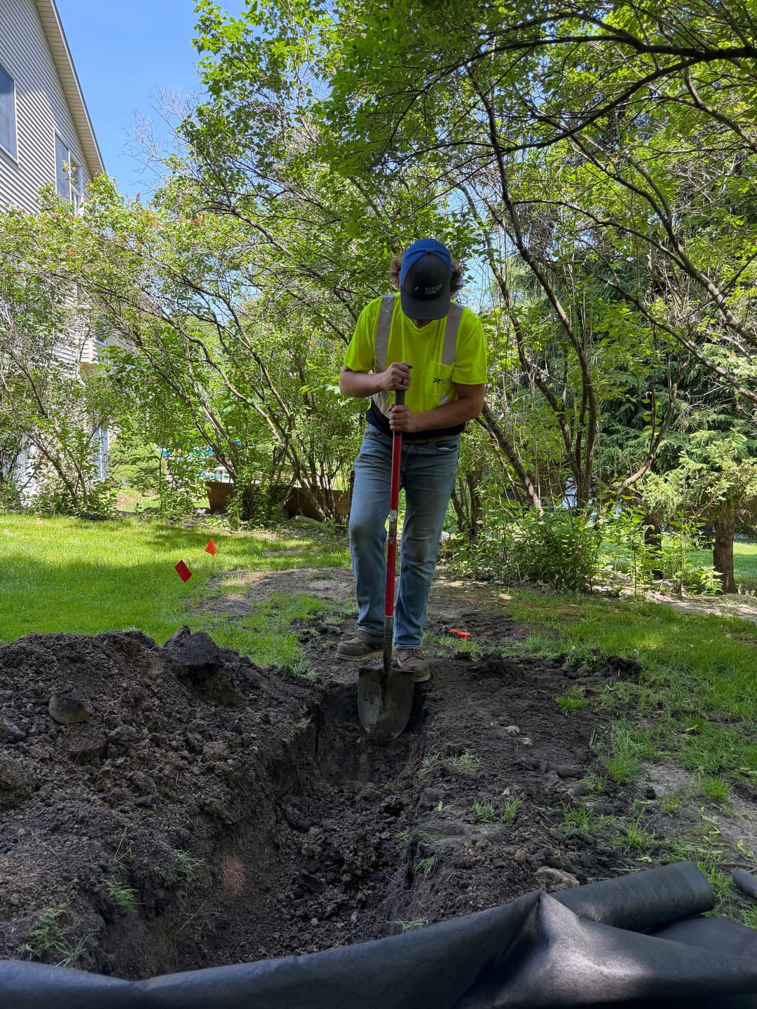 Person digging a trench in a yard with a shovel, surrounded by trees and flags.