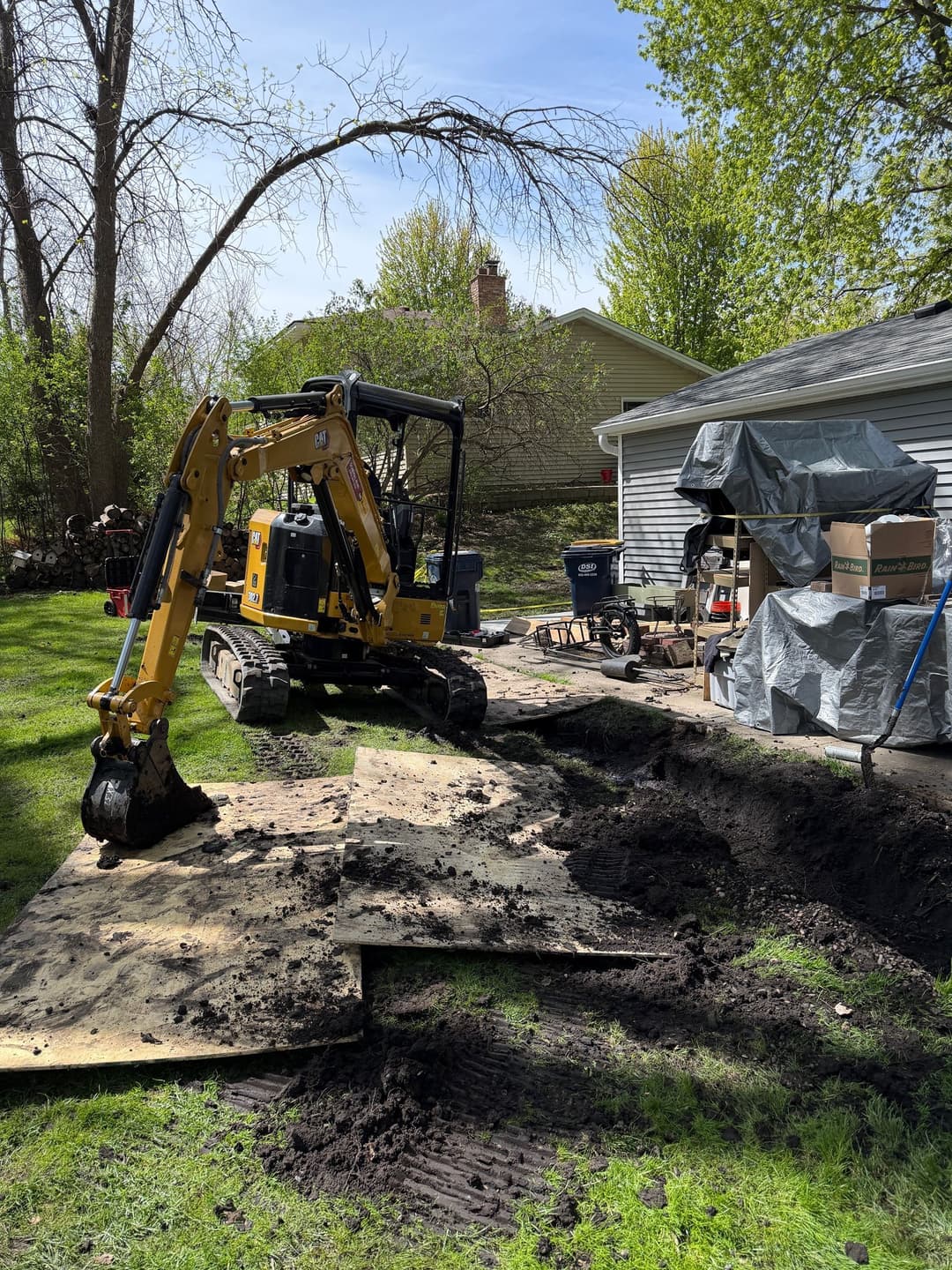 Excavator on residential property, moving soil and debris near house and garden.