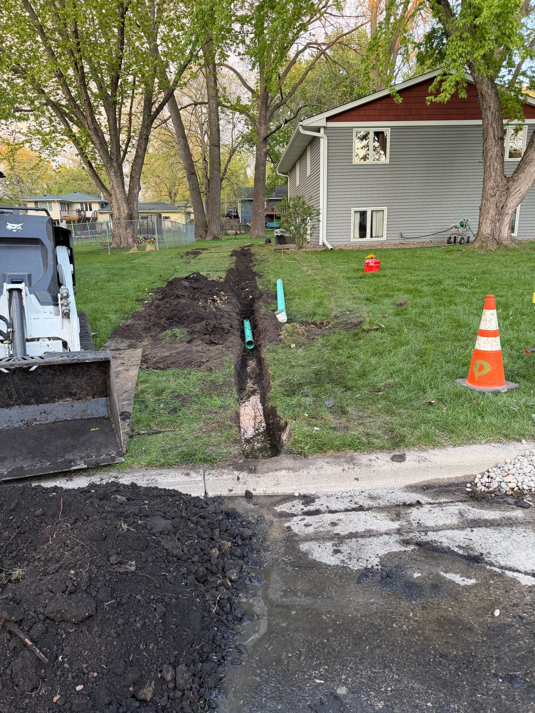 Trenched yard for landscaping with equipment, pipes, and safety cones near a house.