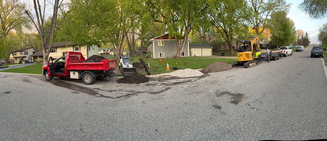Excavation work on residential street with equipment, truck, and landscaping materials.