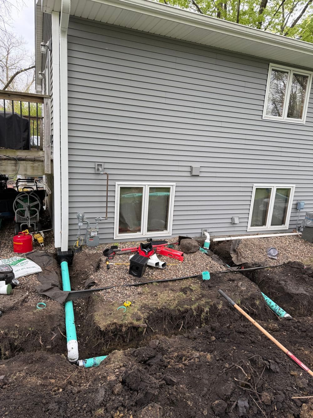 Construction site with excavated soil, PVC pipes, and landscaping tools beside a gray house.