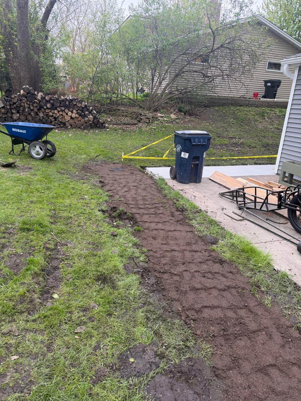 Garden pathway preparation with soil, wheelbarrow, trash can, and stacked firewood.