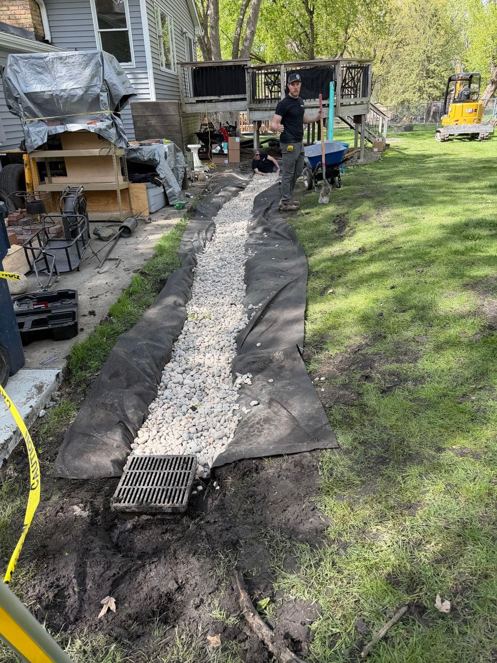 Man installing a gravel drainage path in yard, with tools and machinery nearby.