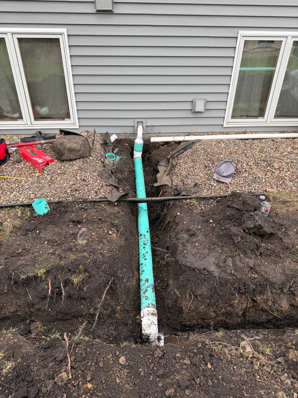 Trenched yard showing blue drainage pipe installation near a house foundation.